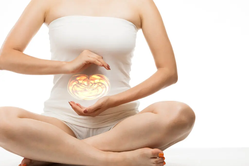 Mulher sentada com as pernas cruzadas, vestindo roupas curtas brancas, e com ambas as mãos em posição de círculo em frente ao intestino, simulando equilíbrio