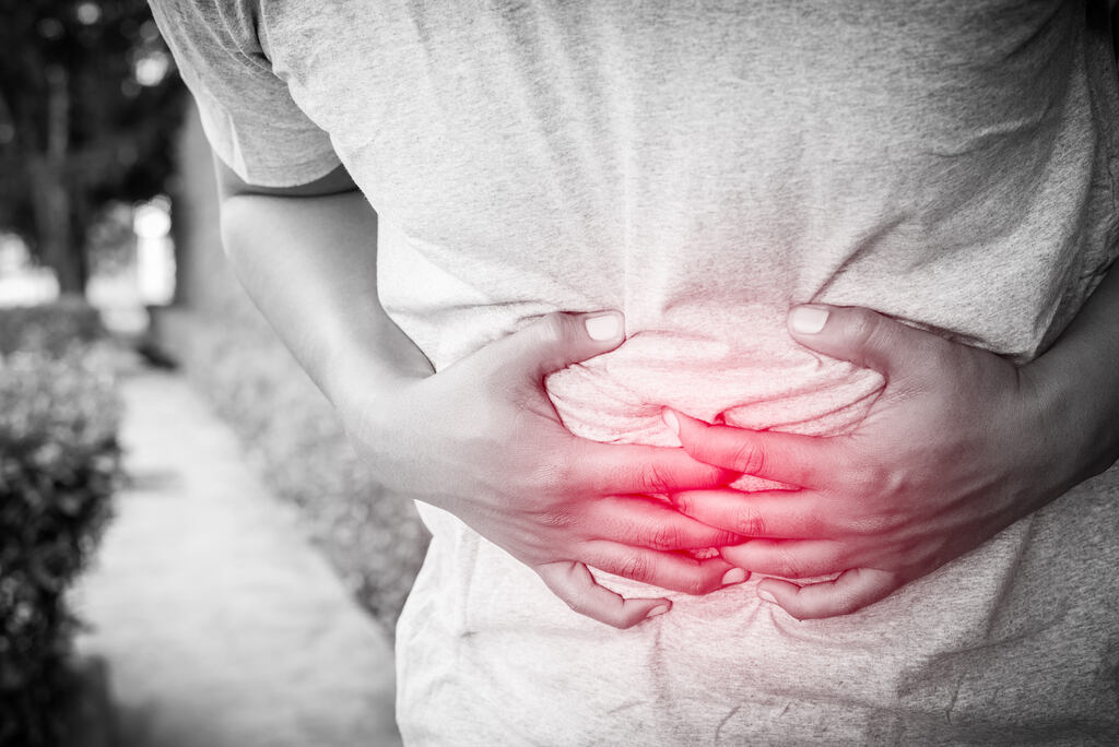 Imagem em tons de cinza, branco e vermelho de pessoa curvada em posição de dor, com ambas as mãos na barriga, simulando dor no instestino