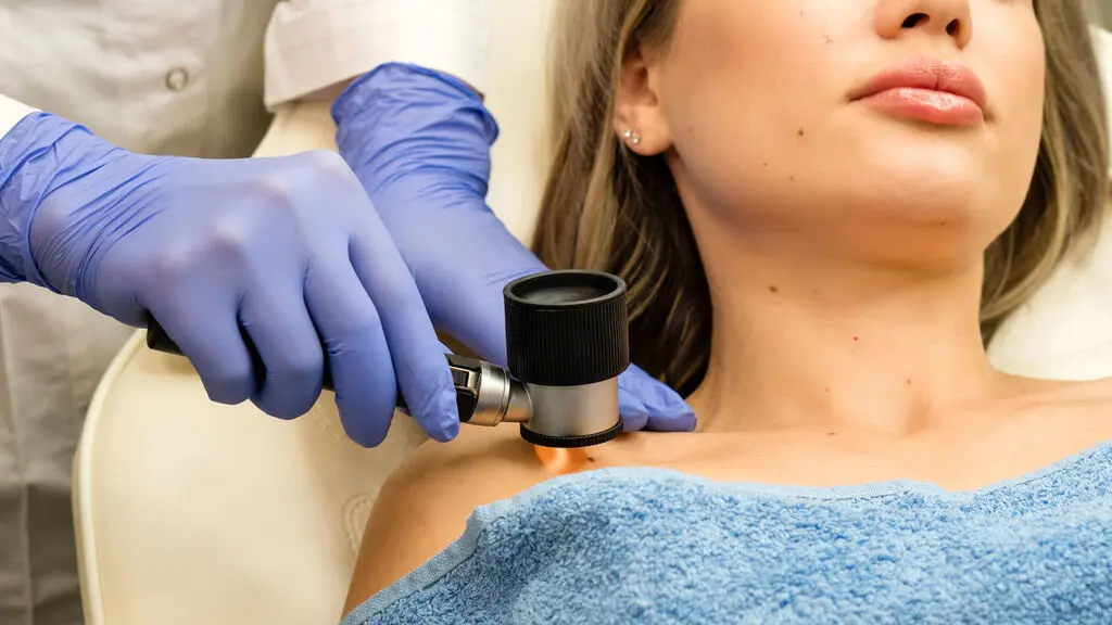 Mãos com luva cirúrgica azul analisam paciente. A mão direita segura um aparelho com luz apontada para a pele da paciente, que tem cabelo loiro longo e está deitada